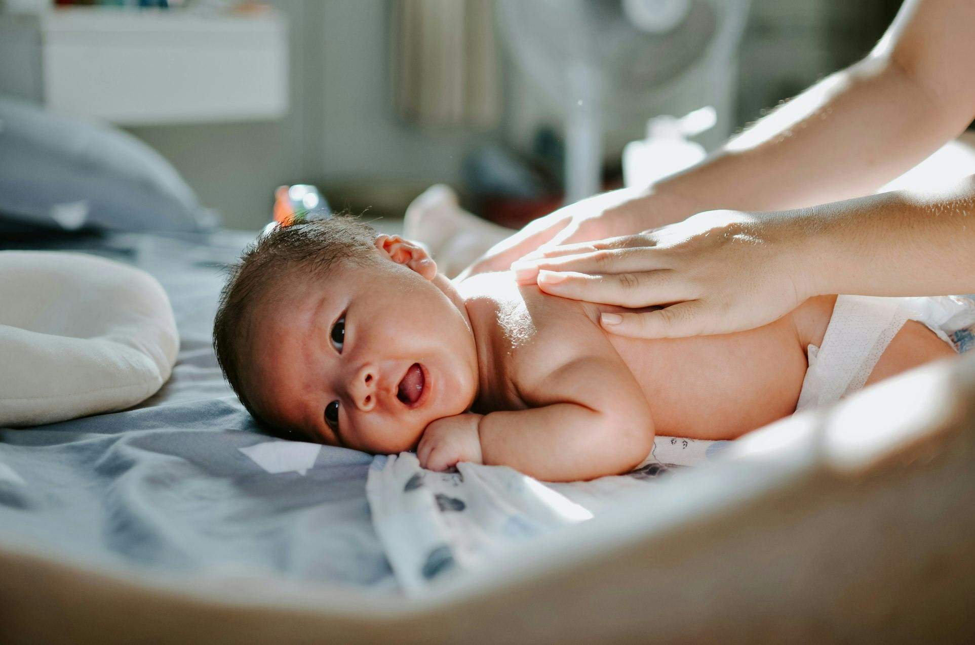 Un nourrisson est allongé sur le côté, regardant attentivement l'appareil photo. Les mains bienveillantes d'un adulte reposent doucement sur le dos du bébé, communiquant tendresse et soin. La lumière naturelle filtre à travers une fenêtre, illuminant la scène d'une douce lumière dorée qui accentue la douceur de l'instant.