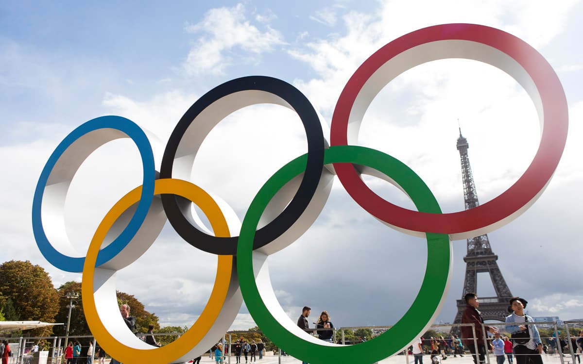 Les anneaux olympiques emblématiques en avant-plan, avec la Tour Eiffel en arrière-plan sous un ciel partiellement nuageux à Paris.