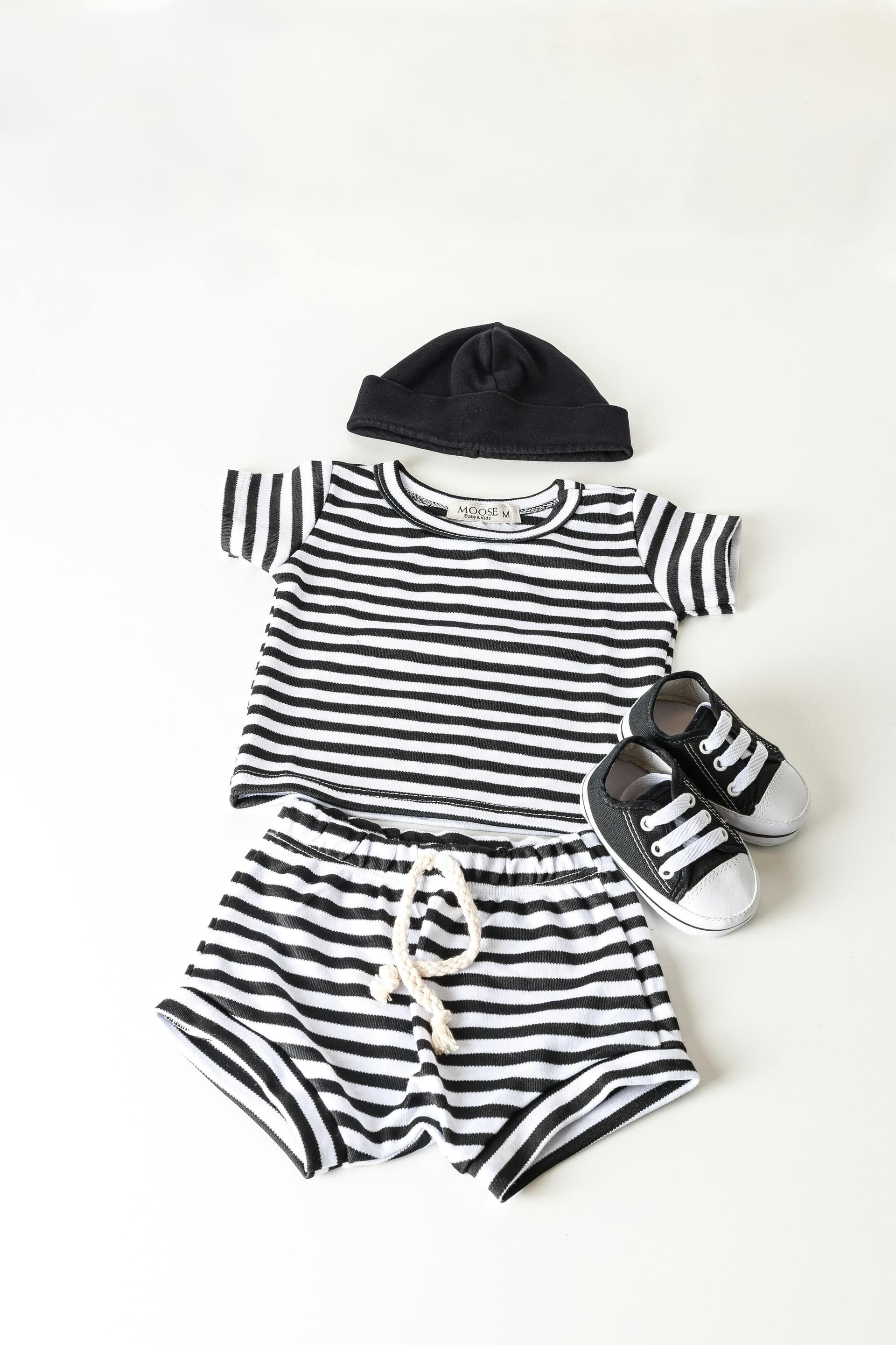 Tenue pour bébé composée d’un t-shirt à rayures noir et blanc, d’un short assorti avec cordon, d’un bonnet noir et d’une paire de petites baskets noires et blanches, disposés sur un fond clair.