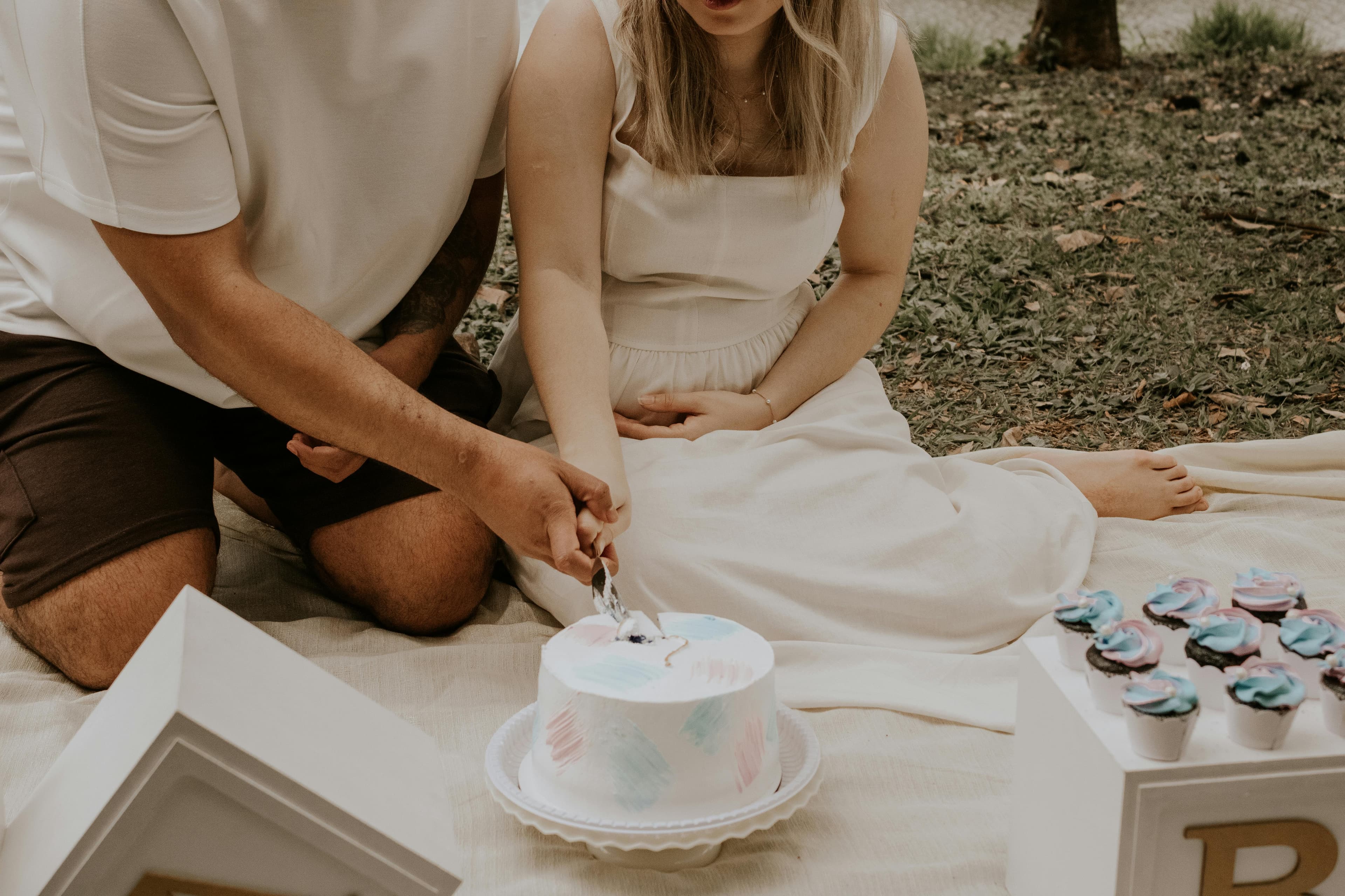 Couple assis sur une couverture en extérieur coupant un gâteau pastel, avec des cupcakes à côté, dans une ambiance douce et naturelle.